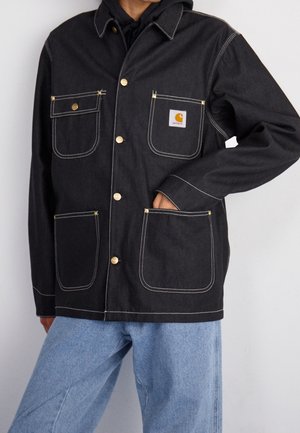 Chaqueta de mezclilla negra con botones dorados y costuras blancas. Presenta dos bolsillos en el pecho, dos bolsillos inferiores y un parche con el logo en el lado izquierdo.