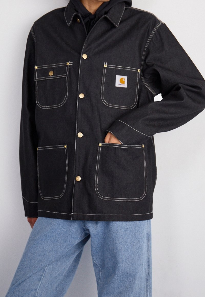 Chaqueta de mezclilla negra con botones dorados y costuras blancas. Presenta dos bolsillos en el pecho, dos bolsillos inferiores y un parche con el logo en el lado izquierdo.