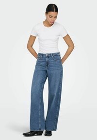 Jean en denim bleu à taille haute et jambes larges avec une finition délavée, associé à un t-shirt blanc ajusté. Bottes noires à talons complètent le look.