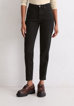 Vrouw draagt hoge taille zwarte rechte jeans en bruine leren veterschoenen, staand tegen een lichte muur.