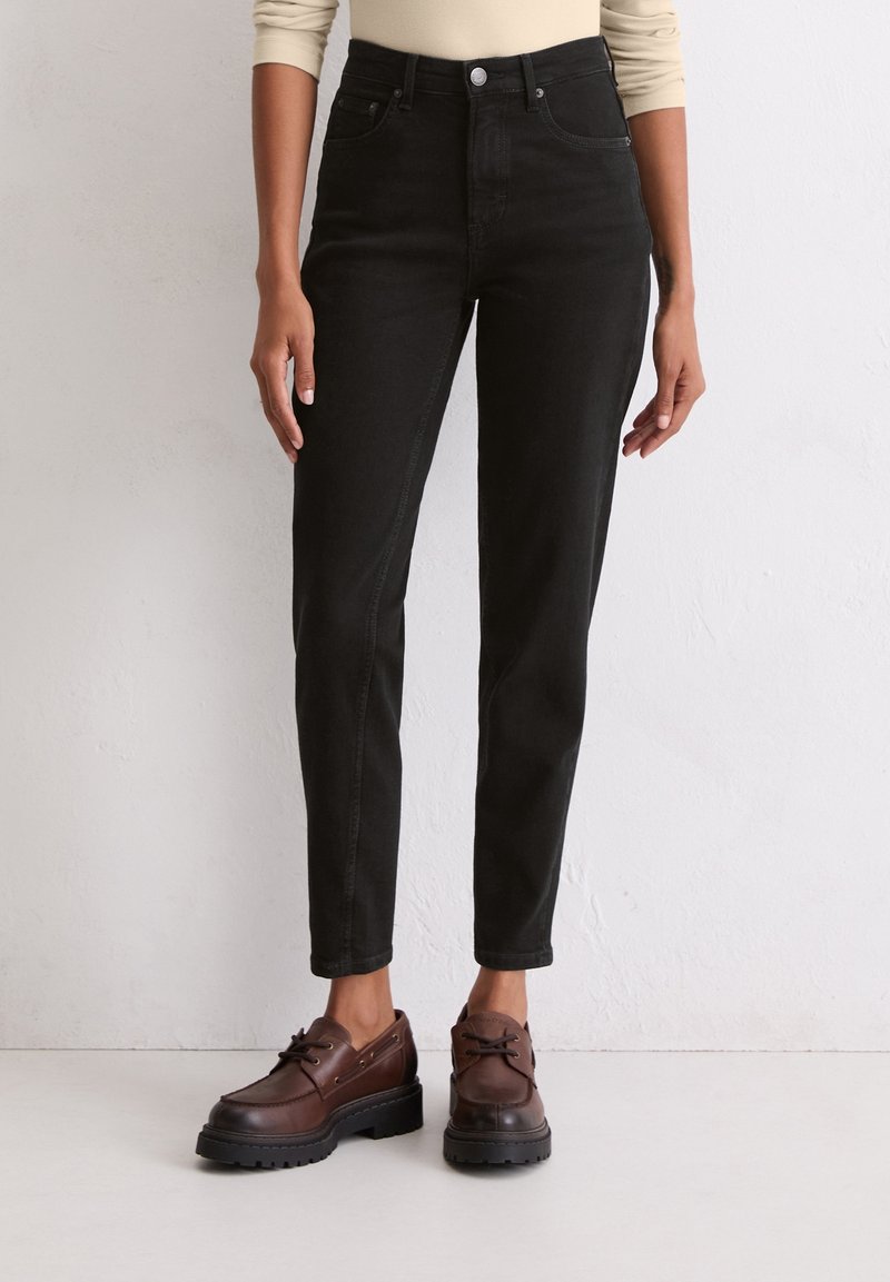 Femme portant un jean noir taille haute à jambes droites et des chaussures en cuir marron à lacets, debout contre un mur clair.