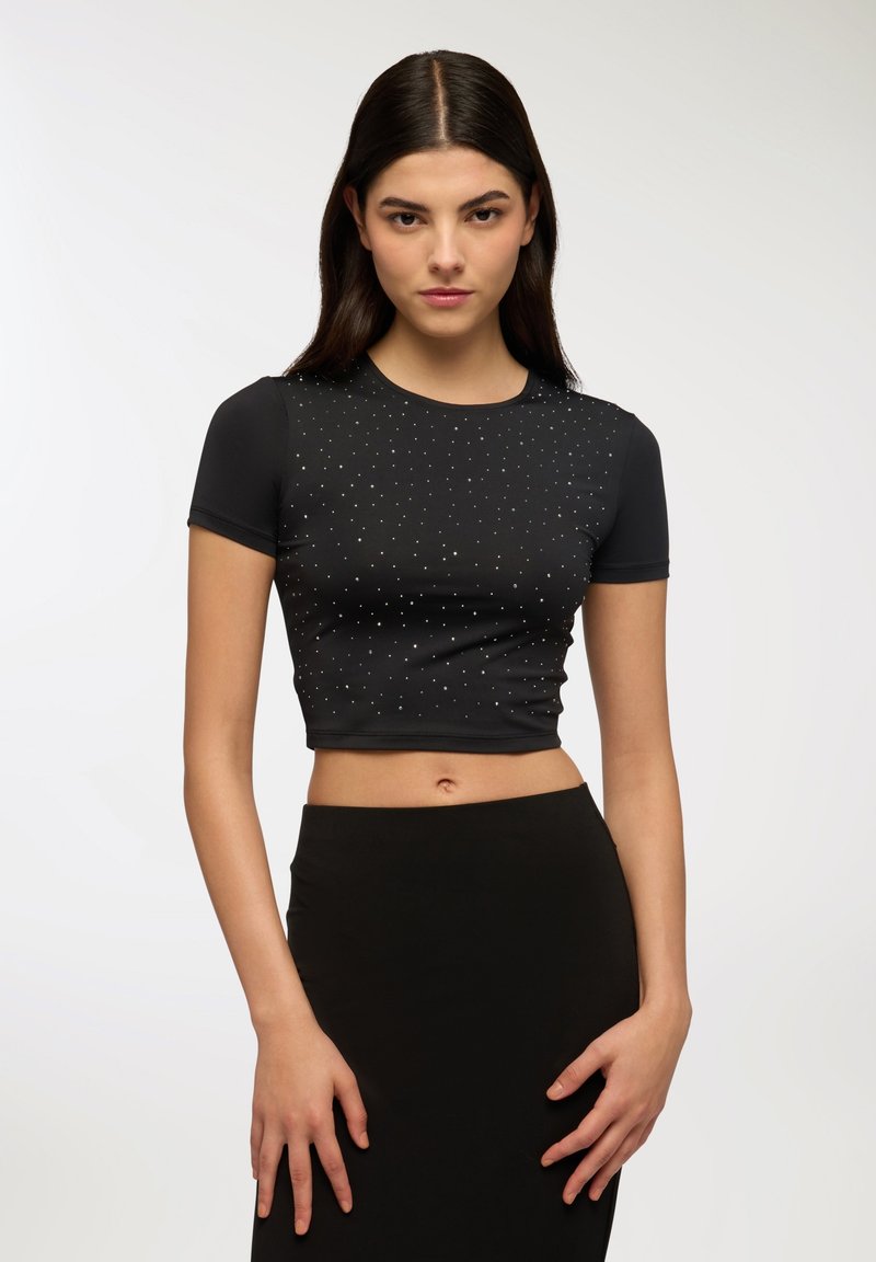 T-shirt nera cropped con maniche corte, decorata con borchie argentate disposte in un motivo, abbinata a una gonna nera. Texture liscia.