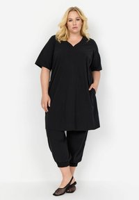 Tunic nera con maniche corte e scollo a V, dotata di due tasche laterali. Abbinata a pantaloni neri con polsini elasticizzati e scarpe slip-on in rete.