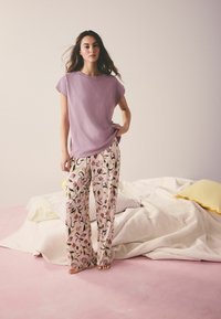 Femme debout pieds nus à côté d'un lit défait, portant un haut à manches courtes lavande et un pantalon large crème avec un imprimé floral.