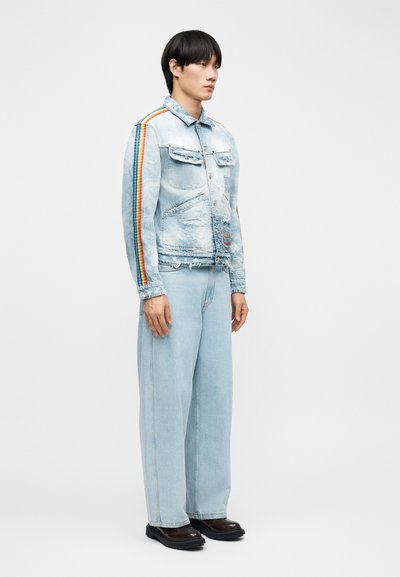 Άνδρας μοντέλο στέκεται φορώντας ανοιχτό γαλάζιο τζιν μπουφάν με ρίγα ουράνιου τόξου στα μανίκια, τζιν παντελόνια φαρδιάς γραμμής που ταιριάζουν και μαύρα παπούτσια.