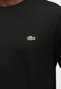 Μαύρο πουκάμισο με στρογγυλή λαιμόκοψη και μικρό πράσινο κεντητό λογότυπο κροκόδειλου στο αριστερό στήθος.