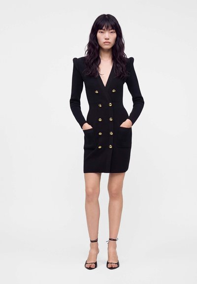 Femme debout portant une robe noire croisée avec des boutons dorés, des manches longues, des épaulettes, des poches et des talons noirs à bride à la cheville.