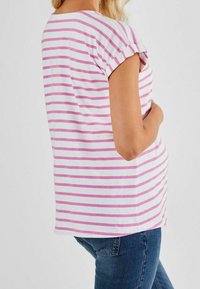 Žena oblečená v bílém a růžovém horizontálně pruhovaném tričku s krátkým rukávem, které doplňuje modrými džíny, stojící na jednobodovém pozadí.