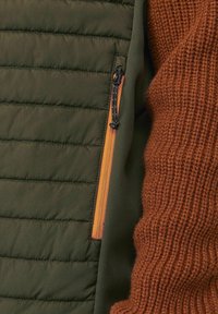 Zielona ocieplana kurtka o pikowanym wzorze z pomarańczowym zamkiem, w zestawie z brązowym swetrem z dzianiny. Gładki materiał kontrastuje z prążkowanym splotem.