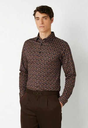 Junger Mann, der ein langärmeliges Hemd mit kleinem Blumenmuster und dunkelbraune Hose trägt, steht mit den Händen in den Taschen.