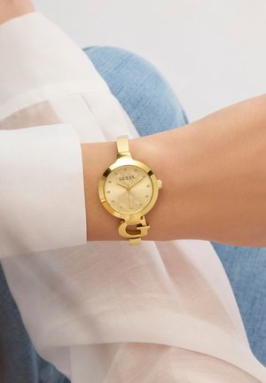 Încheietură purtând un ceas Guess auriu cu marcatori orari din cristale, cadran auriu și detaliu unic în formă de G pe brățară, peste mânecă albă și blugi albaștri.