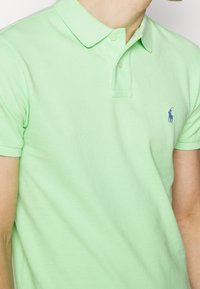 Muž v světle zeleném polo tričku s krátkým rukávem, na levé části hrudi má malé tmavě modré vyšívané logo.