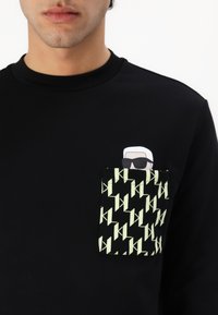 Sort sweatshirt med en tekstureret lomme, der har et gentaget mønster i hvidt og grønt. Pyntet med en lille grafisk figur med solbriller.