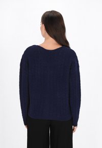 Marineblauwe gebreide trui met een textuur kabelpatroon, oversized pasvorm en een brede V-hals. Lange mouwen reiken tot de polsen.