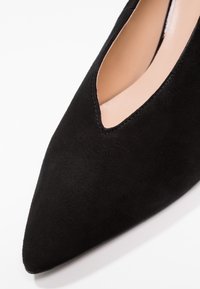 Scarpa nera in suede con punta a becco, con scollatura a V e interno beige su sfondo bianco.