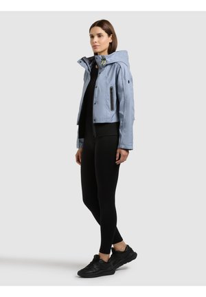 Frau trägt eine hellblaue Kapuzenjacke, schwarze Leggings und schwarze Turnschuhe, steht vor einem einfarbigen hellgrauen Hintergrund.