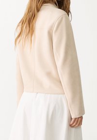 Beżowa kurtka zamszowa ekologiczna z kołnierzem, krótkim krojem i widocznymi szwami, noszona na białej spódnicy.