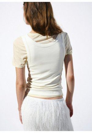 Tânăr văzut din spate purtând o cămașă crem cu mâneci scurte, un top fără mâneci alb și o fustă albă texturată pe un fundal alb.