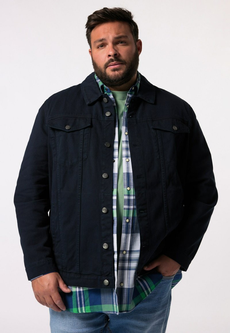 Tmavě modrá džínová bunda s cvoky, s dvěma náprsními kapsami, přehozená přes kostkovanou flanelovou košili v zelené, modré a bílé barvě.