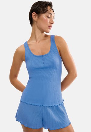 Femme aux cheveux courts et foncés portant un débardeur bleu côtelé sans manches et un short assorti, regardant vers le bas avec les mains derrière le dos.