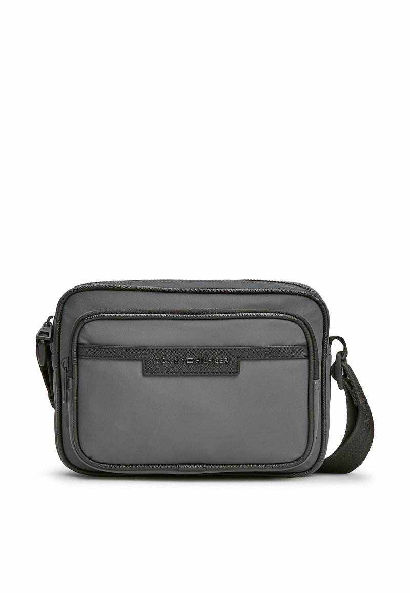 Bolsa mensajera de nailon gris con correa ajustable, que cuenta con un bolsillo frontal con cremallera y un parche de logo texturizado. Diseño compacto y rectangular.