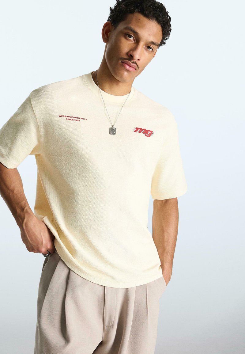 Jeune homme portant une chemise à manches courtes crème avec un texte rouge, un pantalon beige et un collier, debout avec une main dans la poche.