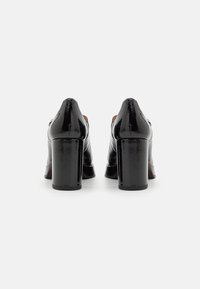 Chaussures à talons block en cuir verni noir avec une finition lisse, un bout arrondi et des détails de couture visibles sur les coutures.