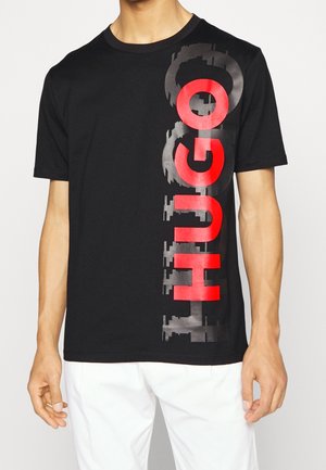 Schwarzes T-Shirt mit kurzen Ärmeln und großem vertikalem roten und grauem "HUGO" Text auf der Vorderseite, getragen mit weißen Hosen von einer stehenden Person.