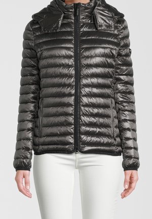 Doudoune noire matelassée zippée avec pattes d'épaule à boutons-pression portée par une personne en pantalon blanc, visible du cou aux hanches.
