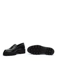 Czarne skórzane mokasyny z srebrnymi ćwiekami, masywna podeszwa z bieżnikiem; jeden but w pozycji pionowej, a drugi pokazujący podeszwę z wytłoczonym logo marki.
