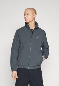Chaqueta ligera de color gris oscuro para hombre, con cierre de cremallera, cuello alzado y puños elásticos, y un pequeño logo en el pecho.