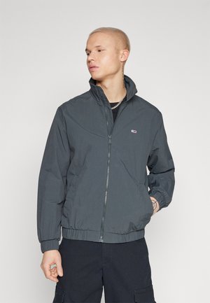 Dunkelgraue Herren-Zipjacke aus leichtem Material, mit Stehkragen und elastischen Bündchen, versehen mit einem kleinen Logo auf der Brust.