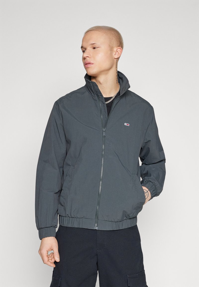 Chaqueta ligera de color gris oscuro para hombre, con cierre de cremallera, cuello alzado y puños elásticos, y un pequeño logo en el pecho.