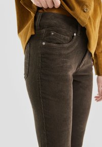 Braune Cord-Hosen mit vertikalen Rippungen, Fünf-Pocket-Design, Metallbeschläge, kombiniert mit einem senfgefärbten, strukturierten Hemd.