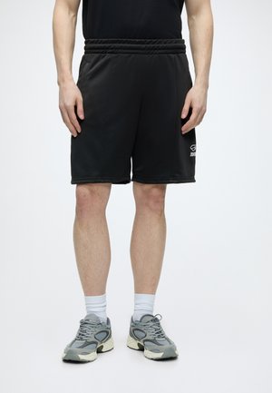 Uomo che indossa pantaloncini sportivi neri, calzini bianchi con risvolto e scarpe da corsa grigie, in piedi con le mani rilassate lungo i fianchi su sfondo bianco.