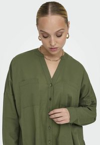Chemise olive verte à col en V, avec deux poches poitrine et des manches longues. Tissu lisse et léger avec une coupe décontractée.