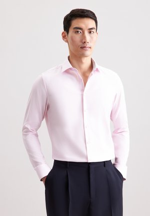 Helles rosa Hemd mit langen Ärmeln, weicher Textur und kontrastierendem gemustertem Kragen. Getragen mit dunklen, maßgeschneiderten Hosen.