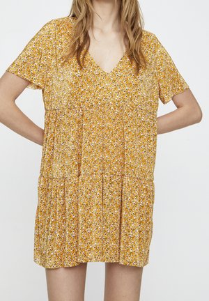Frau trägt ein locker sitzendes, kurzärmliges, gelbes Kleid mit kleinem weißem Blumenmuster und V-Ausschnitt, steht mit den Händen in der Hüfte.