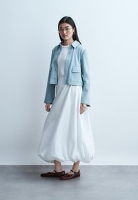 Moteris su ilgais tamsiais plaukais ir akiniais, vilkinti baltą su balionais apačioje suknele, šviesiai mėlyną užsegamą švarką ir rudas Mary Jane tipo basutes, stovinti ant pilko grindinio.