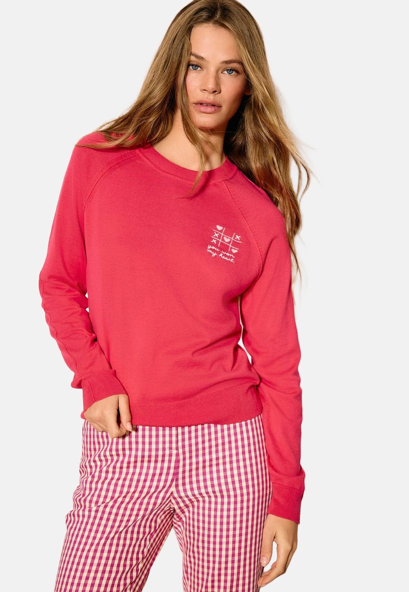 Sweat-shirt rose à manches longues avec des poignets côtelés, présentant un petit motif graphique blanc sur la poitrine. Assorti à un pantalon à carreaux rose et blanc.