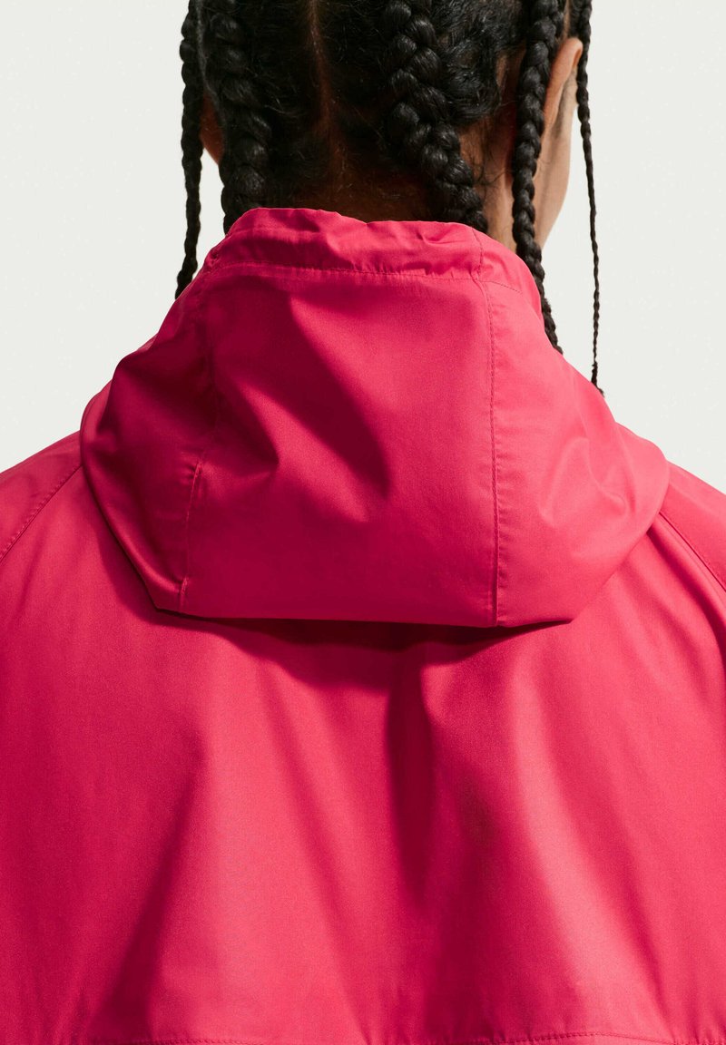 Persoon met donker gevlochten haar die een felroze capuchonjack draagt, gezien van achteren tegen een effen lichte achtergrond.