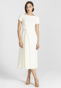 Kremowa sukienka za kolano z krótkimi rękawami, z charakterystycznym skręconym detalem w talii i zwiewnym dołem. Gładki materiał o miękkiej fakturze.