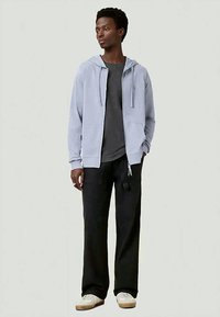 Fleeced de capuz azul claro com fecho éclair e cordões, usado por cima de uma t-shirt cinza escura, combinado com calças cargo largas pretas e ténis bege.