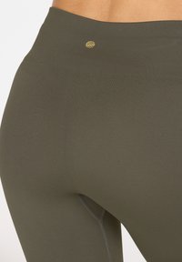 Hoch taillierte Leggings in dunkelolivgrün, aus elastischem Stoff gefertigt. Mit einem kleinen goldenen Logo am Bund und Flachnähte.