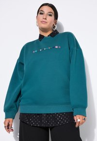 Felpa color turchese con motivi di gemme colorate ricamati sul petto, indossata sopra una maglietta nera con piccoli motivi bianchi.