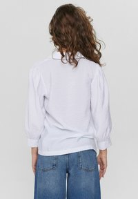 Blusa bianca con texture, colletto, maniche a palloncino e orlo curvo. Abbinata a jeans a gamba larga in denim blu. Vista da dietro.