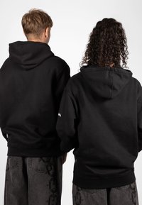 Zwarte hoodies met een relaxte pasvorm, gemaakt van een katoenmix. De achterzijde toont stikdetails en een logo op één mouw. Donkergrijze broek met een kettingpatroon.