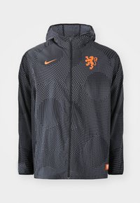 Nike jakna u crnoj boji s točkastim uzorkom, s narančastim grbom lava i logotipom. Uključuje kapuljaču i zatvaranje s patentnim zatvaračem.