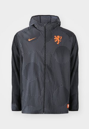 Nike jacket in black with dotted pattern, featuring an orange lion emblem and logo. Includes a hood and zippered front.