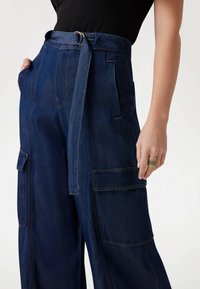 Pantaloni cargo ampi in denim scuro con grandi tasche laterali e vita con cintura. Il tessuto presenta cuciture visibili e una texture morbida.